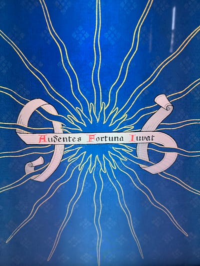 The image shows a blue and yellow banner or flag with a stylized design. In the center is a vertical ribbon with the Latin phrase "Audentes Fortuna Iuvat" written in red and black lettering. The ribbon curls at both ends, resembling a scroll. Surrounding the ribbon are numerous thin, yellow rays emanating from a central point, creating a sunburst effect. The background is a textured blue with a repeating pattern that is difficult to discern, although the pattern is made up of squares.

Based on the motto "Audentes Fortuna Iuvat" (Fortune favors the bold), this design is likely associated with the city of Katwijk, Netherlands. The motto is found on the Katwijk coat of arms.