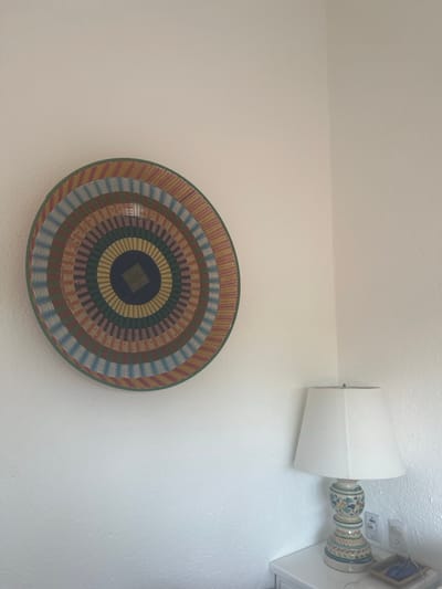 A woven, circular piece of art hangs on a white wall. It features concentric rings of colors, including brown, blue, green, and yellow, emanating from a dark blue square at the center. Above it and to the right, a table lamp is mounted on the wall, with a white lampshade and a decorative ceramic base in shades of white, green, blue and brown. The background is a plain white wall.