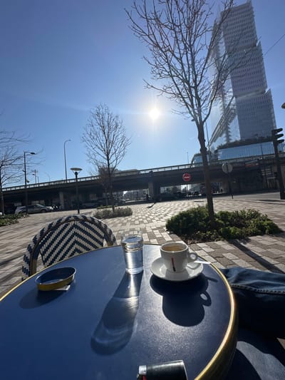 The image presents a sunlit outdoor cafe scene in Clichy, France, with the Tours Duo skyscrapers visible in the background. In the foreground, a dark blue circular cafe table with a gold trim holds a white espresso cup and saucer with a red vertical logo, a clear glass of water, and a blue, empty ashtray. A black lighter rests near the bottom edge. A person's knee, covered in blue denim jeans, is visible at the lower right. To the left, a portion of a cafe chair featuring a blue and white geometric woven pattern is present.

The immediate environment consists of a paved pedestrian area with small, dark green bushes and several bare deciduous trees casting shadows. An elevated concrete road or overpass traverses the midground horizontally, supporting what appear to be traffic lanes. Below this overpass, a street with several parked and moving vehicles is visible, along with a red "no entry" sign. Beyond the overpass and to the right, the distinctive, multi-faceted glass towers of Tours Duo rise prominently against a clear, bright blue sky. The sun is intensely bright in the upper mid-right, creating a strong glare and casting sharp shadows across the scene. Streetlights and traffic signals are also visible.