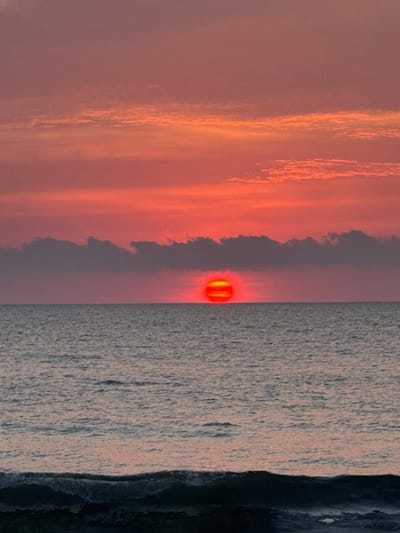 An orange sunset dominates the sky, with fiery hues radiating from a low-hanging sun. Wispy clouds streak the sky, fading into softer, muted tones. The sun's glow reflects faintly on the surface of the water, which extends out to a dark coastline on the right.