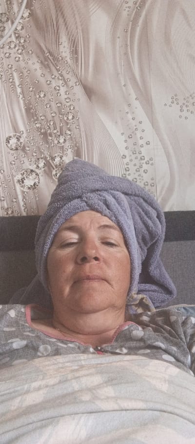 An overhead, close-up shot shows a woman lying down with a purple towel wrapped around her head. Her eyes are closed, and she appears to be resting. She is wearing a grey and white polka dot pajama top with a pink trim. The background shows a textured wallpaper with a pattern of diamonds and flowing lines. The woman is lying on what appears to be a bed or couch. The image is taken indoors. The location in the city of Beregovoe, Ukraine is not discernible from the image.