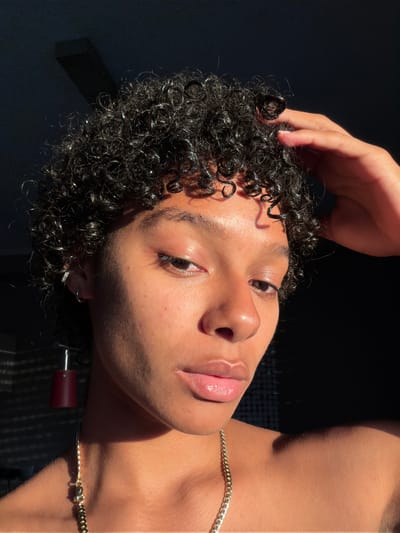 A close-up, frontal view shows the upper body of a person with dark, curly hair, touching their head with their right hand. The person has light brown skin and is wearing a gold chain necklace and a silver hoop earring in their left ear. A red cylindrical earring hangs from their left earlobe. The lighting suggests natural light is coming from the left, casting shadows across the person's face and body. The background is dark and out of focus, with faint vertical lines visible on the left side, possibly indicating blinds or a window. There is no discernible location in Champs-sur-Marne, France.