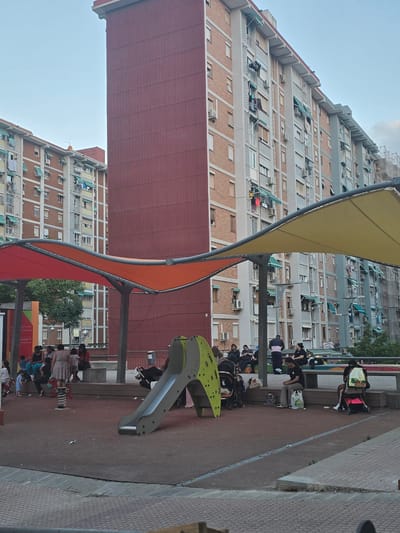 The image depicts a playground area in front of residential apartment buildings in Cornellà de Llobregat, Spain. The buildings are multi-story, with balconies and visible air conditioning units. In the foreground, a playground structure with a slide and a red spring rider is visible. Several people, including adults and children, are present in the playground area, some seated on benches, others standing or pushing strollers. Colorful fabric canopies extend over part of the playground, providing shade. The ground of the playground appears to be a dark, rubberized surface. Trash is scattered on the ground in the playground area.
