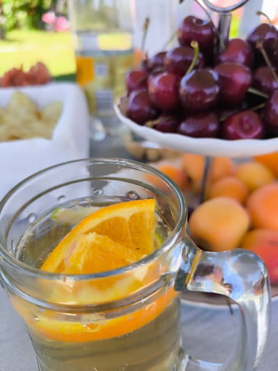 A glass of water with orange slices sits on a table, with cherries and apricots in the background. The scene appears to be outdoors, with green grass visible in the background. The location is not specified in the image.