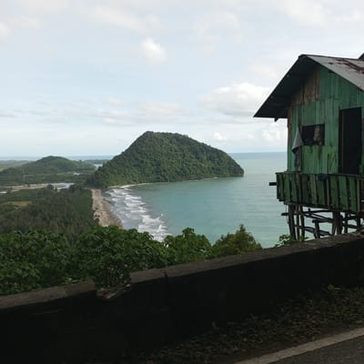 This image captures a coastal view in Meudheun, Indonesia. The scene is dominated by lush green hills sloping down to a sandy beach, where waves gently roll onto the shore. The turquoise ocean stretches out to the horizon, meeting a pale sky with scattered clouds.

In the foreground, a rustic wooden house, painted a faded green, stands on stilts overlooking the sea. The house has a corrugated metal roof and a small balcony with a wooden railing. A white cloth and some other items are visible hanging from the house. The house appears weathered, with visible signs of age and disrepair.

A dark, textured concrete barrier runs along the bottom of the image, suggesting a road or elevated vantage point. The surrounding vegetation consists of dense green trees and foliage, some of which are in the immediate foreground. The overall impression is one of a remote, natural setting with a simple dwelling integrated into the landscape.