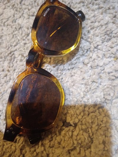 The image shows a pair of MOSCOT sunglasses on a textured surface, likely carpet. The glasses are predominantly amber and tortoise shell in color. Light reflects off the lenses, giving them a warm, brown hue. The frame is clearly visible, and the MOSCOT logo is subtly displayed near one lens. The lighting creates a shadow behind the glasses, adding depth to the image. The texture of the carpet provides a soft backdrop, contrasting with the smooth, glossy surface of the sunglasses.