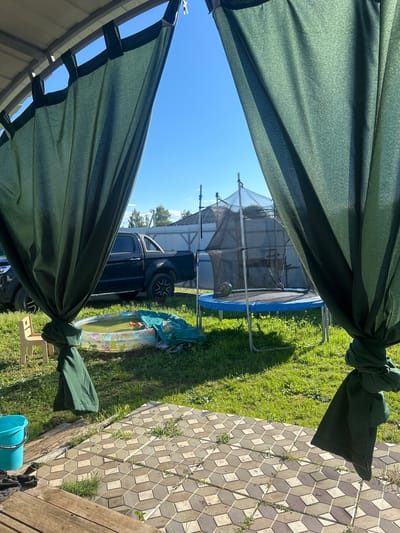 The image depicts a backyard scene on a sunny day. In the foreground, a dark green canopy or tarp is draped, obscuring a portion of the view. Through the gap in the canopy, a backyard is visible, characterized by lush green grass.
Prominently featured is a trampoline with a black safety net and a blue mat. To the right of the trampoline, a dark-colored pickup truck is parked. Next to the truck, a small inflatable pool is visible, containing water and what appears to be a toy. A light blue bucket is also present in the lower right corner of the image, along with some wooden planks that suggest a deck or patio. The ground is partially covered with patterned square tiles.
The sky is clear and blue, indicating fair weather. The lighting suggests it is daytime. The overall scene conveys a sense of leisure and outdoor activity, possibly a family enjoying their yard. No people are visible in the frame. No text is discernible in the image. The location context provided is Лотошино, Russia.