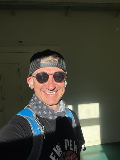 A man is captured in a selfie, standing indoors with a door visible in the background to his left. He is wearing a black baseball cap, sunglasses, a grey and black patterned neck gaiter, and a black t-shirt. A blue and grey backpack is visible on his shoulders, with a keychain hanging from one of the straps. The lighting suggests a sunny day, with shadows cast on the wall and floor, indicating the presence of windows. The environment appears to be a functional space, possibly a school or public building, with overhead conduits visible in the upper portion of the frame. The man is smiling, looking directly at the camera. The location is identified as Bellelay, Switzerland.