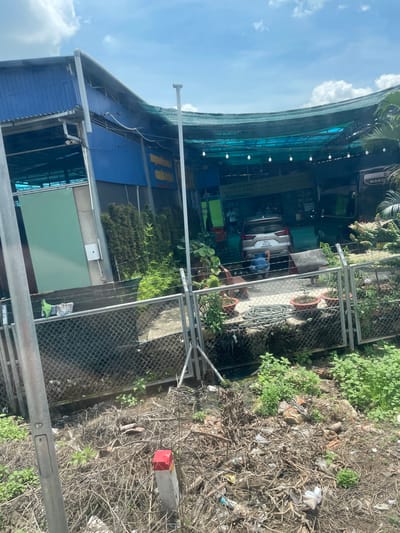 The image shows a car wash in Binh Xuan 2, Vietnam. A silver SUV is parked in the bay, and a person is kneeling beside it, appearing to wash the car. The car wash has a blue corrugated metal facade and a green tarp roof stretched over a metal frame, supported by posts. String lights are hung under the tarp, illuminating the area. A sign with yellow lettering is visible on the back wall of the car wash. The facility is bordered by a chain-link fence. Outside the fence, the ground is overgrown with weeds and littered with debris, including plastic bottles, branches, and other trash. A grey metal pole is positioned in the foreground, and a concrete marker with a red top is visible on the right side of the frame amidst the vegetation and debris. The sky above is blue with scattered white clouds.