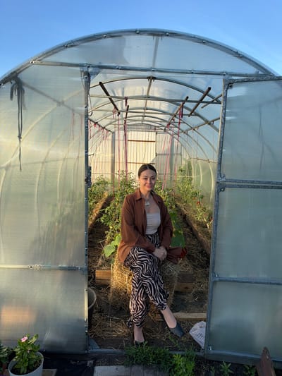 A full-shot, outdoor photograph captures a woman sitting on a bale of hay inside a greenhouse. The greenhouse has a transparent, corrugated polycarbonate roof supported by a metal frame. The woman is positioned in the center of the greenhouse, facing the camera. She has dark hair pulled back and is wearing a brown button-down shirt over a light-colored tube top, and black and white zebra-patterned pants. She is also wearing black slip-on shoes. Her hands are resting on her lap.
Inside the greenhouse, rows of plants, appearing to be tomatoes and cucumbers, are growing. The ground is covered with straw and some wooden planks. A white bucket is visible to the left of the woman. Outside the greenhouse, to the lower left, a potted pink flower is visible. The sky above is a clear blue, suggesting it is daytime, likely late afternoon or early evening due to the warm lighting. The scene appears to be in a rural or suburban setting. There is no information to suggest this location is in Kansk, Russia.