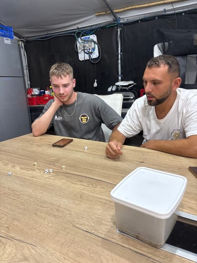 The image portrays two males seated at a light-colored wooden-grain table in an indoor setting in Mazkeret Batya, Israel. Six white dice are scattered across the table surface. The male on the left, with short light-brown hair, wears a grey t-shirt displaying a BAPE ape logo and "A.D.J." text, along with a silver chain. He is seated, resting his left elbow on the table and supporting his chin with his left hand, looking down at the dice. A brown smartphone lies face down near his position on the table. The male on the right, with short dark hair and stubble, wears a white t-shirt featuring a partially visible "Hurley Surf" logo. He is seated, leaning forward, with his right hand visible on the table near a translucent rectangular plastic container with a white lid. He observes the dice on the table.
In the background, to the left, a silver-grey refrigerator is visible, topped with a blue plastic crate labeled "אריזה" (packaging) in Hebrew. A red plastic crate containing various items is positioned behind the left male. The rear wall consists of a dark, possibly fabric or tarp-like material. A white panel, labeled "אקסטרא" (extra) in Hebrew, equipped with multiple hoses and gauges, is mounted on this wall. The ceiling is light-colored, with exposed conduits secured by blue ties.