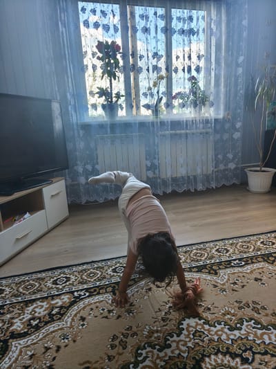 The image depicts a young child in a downward dog yoga pose on a patterned rug. The child is wearing light-colored leggings and a pink t-shirt. Their head is down, and their hands are on the rug. A doll with reddish-brown hair lies on the rug near the child's hands. In the background, a television set is visible on a wooden stand to the left. To the right, a window with white lace curtains is open, revealing plants on the windowsill and the sky outside. A potted plant stands to the far right. The flooring appears to be light-colored wood laminate. The scene is indoors, consistent with a residential setting.