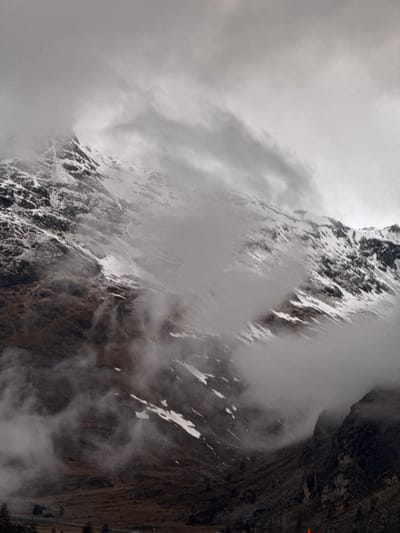 The image captures a dramatic alpine landscape, dominated by a towering mountain or cliff face partially shrouded in swirling clouds and mist. The main subject is a rugged, steep rock formation, visible on the left side, characterized by jagged textures and patches of white, likely snow or ice, clinging to its surfaces. The atmospheric conditions are overcast and misty, diffusing the light and creating a soft, ethereal quality throughout the scene.
On the right side of the image, a darker, imposing rock wall descends vertically. Along the lower right edge of this cliff, a narrow path or road is discernible, appearing to be carved into the mountainside. A small, dark-clad human figure is visible on this path, suggesting an individual traversing or observing the scene. Guardrails or barriers appear to line the path, indicating a developed route for travel.
The weather is distinctly cloudy and misty, likely contributing to a cool or cold environment given the snow. There is no clear indication of a specific time of day beyond daylight, but the diffused light suggests an overcast sky rather than direct sunlight. The scene evokes a sense of grandeur, scale, and the adventurous spirit of exploring high-altitude terrain. The location context of Pontresina, Switzerland, aligns with the appearance of a majestic alpine setting. There is no visible text in the image.