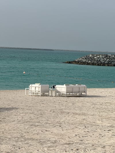 The image shows a beach scene in what is likely Ghadeer Al Tayr, United Arab Emirates. The view is oriented so that the sky is on the left and the sandy beach is on the right. In the foreground are several white beach chairs and what appear to be cabanas or sunbeds. The middle ground is dominated by the turquoise sea, which has a line of yellow buoys marking a boundary. A rocky breakwater extends into the sea on the left side of the frame. The sky is overcast. No people are visible in the image.