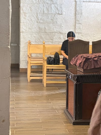 A woman in a black t-shirt and black pants is sitting on a wooden bench in a building. She is looking down at her phone. There are two black bags on the bench next to her. The bench is in front of a wall covered in light-colored stone tiles. To the right of the bench is a dark wooden podium covered with a burgundy cloth. The floor is tiled with light brown wood-look tiles. The scene appears to be inside a church or a similar public building. The city of Masis, Armenia is not visible in the image.