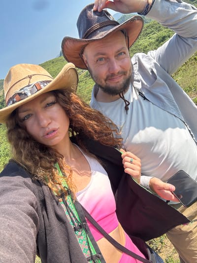 A selfie shows a woman and a man in a natural landscape in Bắc Hà, Vietnam. The woman, wearing a cowboy hat with a bull motif, is positioned below the man. She is wearing a white and pink top under a dark shirt with a patterned trim. Her hair is curly and auburn. The man, above her, also wears a cowboy hat. He is holding a black mobile phone. He has a beard. He is wearing a light colored shirt. The background consists of a green mountainous terrain. The sky is visible.