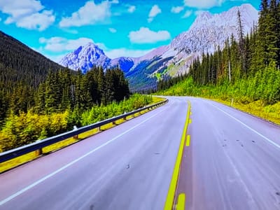 The image shows a paved road with yellow and white lane markings winding through a mountainous, forested landscape. The road is bordered by a guardrail on the left and dense evergreen trees on the right. In the distance, majestic, snow-capped mountains rise against a bright blue sky with scattered white clouds. The overall scene depicts a scenic natural environment, likely a highway passing through a national park or mountainous region. The location is not in Gorzow, Poland, as Gorzow is a city with a predominantly flat landscape and is not characterized by the mountainous terrain depicted in this image.