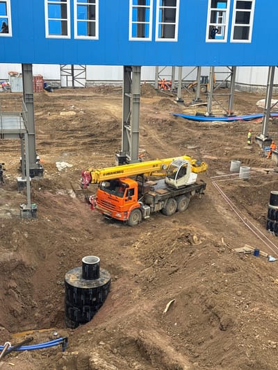 This is a top-down, rotated image of a construction site in Chulman, Russia. The scene features a large, blue building with several windows on the left side, and a construction area on the right. The construction area is characterized by exposed earth, mounds of dirt, and what appear to be pipes and construction materials.

Prominently featured is a yellow and orange crane truck, with visible text that appears to read "ИВАНОВЕЦ" (Ivanovets) and "KAMAZ" on the truck's cab. The crane is in a lowered position. Several construction workers, wearing orange and yellow vests and hard hats, are scattered throughout the site, engaged in various tasks. One worker is visible in an open window of the blue building, and another appears to be inside the crane's cab.

In the background, other construction equipment, including a yellow bulldozer, is visible. There are also large concrete pipes and coils of black material, possibly insulation or conduit, embedded in the ground. Blue pipes are also visible, running along the ground. The ground surface is uneven and appears to be composed of dirt and gravel, with some areas showing signs of recent excavation. The lighting suggests it is daytime, with no clear indicators of weather conditions. The overall impression is one of active construction and industrial activity.