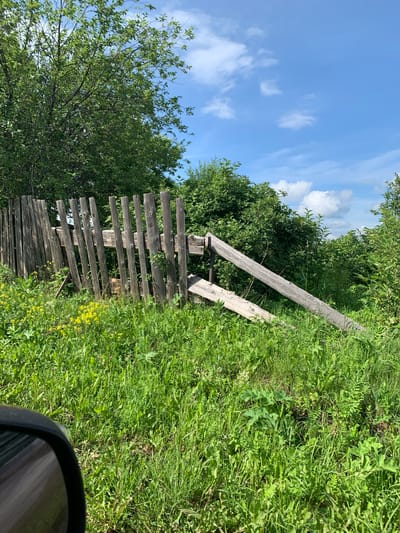 The image depicts a broken wooden fence in a rural setting, likely in the city of Barsai, Russia. The fence, made of vertical wooden planks, is leaning and appears to be in disrepair, with one section significantly collapsed. It is surrounded by lush green grass and vegetation, including some yellow wildflowers. In the background, dense green foliage and trees are visible under a bright blue sky with scattered white clouds. The lower left corner of the image shows the reflection of a car's side mirror, indicating the photo was taken from a vehicle. No people are present in the scene. The environment suggests a summer day in a countryside location.