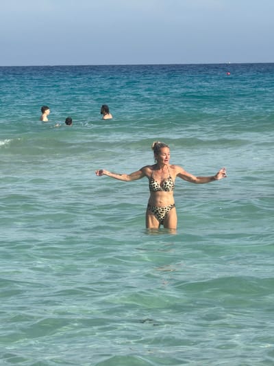 An overhead shot captures a woman floating on her back in the clear, shallow waters of the sea, near Ayia Napa, Cyprus, on a sunny day. She is wearing a leopard print bikini, with her arms slightly raised and head tilted back, indicating relaxation. Her body is partially submerged, showing her torso and legs below the water's surface.

Further out in the deeper, bluer water, the heads of three other individuals are visible, also swimming or standing in the sea. The water ranges from a light, translucent turquoise where the main subject is, to a deeper blue towards the horizon. The sky above is a clear, light blue. In the far distance, a small red buoy can be seen on the water's surface. No visible text is present in the image.
