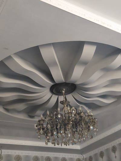 The image shows a chandelier hanging from a ceiling. The ceiling has a decorative, wavy design. The chandelier is ornate and appears to be made of metal and crystal. The room is lit by the chandelier, and the walls have decorative wallpaper. The scene is likely an interior shot of a residential or commercial building. There is no visible indication of the city of Preobrazhenskoye, Russia.