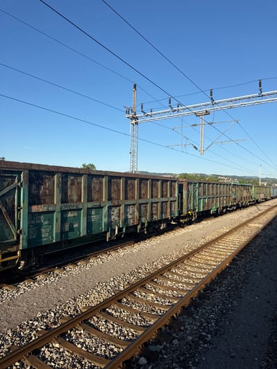The image shows a train of open-top freight cars, likely used for transporting bulk materials like coal or gravel, on a sunny day. The train is stationary on a railway track. The cars are old and appear weathered, with rust and graffiti visible on their surfaces. The railway line is surrounded by ballast, and in the background, there are trees and a city skyline, suggesting an urban or semi-urban setting. Overhead power lines and a supporting pylon are also visible, indicating an electrified railway. The scene is viewed from a slightly elevated position, looking down the length of the train and track. The sky is clear and blue. Without further information, the specific location within Valjevo, Serbia cannot be determined from the image alone, but the presence of railway infrastructure and a city in the background suggests it is likely situated near or within the city limits.
