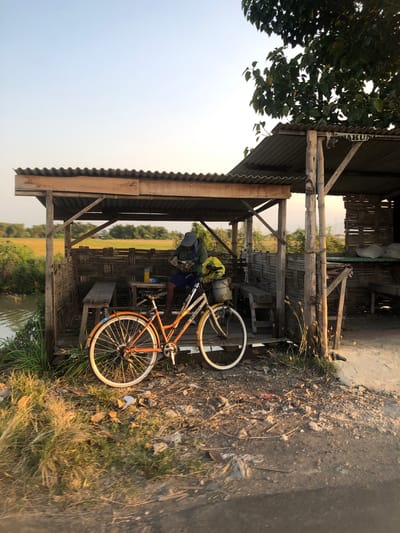 The image depicts a humble, open-air shelter constructed with corrugated metal roofing and bamboo woven walls, situated near a body of water and a rural landscape, possibly a rice paddy field, indicative of a location in Keboguyang, Indonesia. A man, wearing a blue baseball cap and a green long-sleeved shirt, is seated within the shelter, appearing to engage with something in his hands. Beside him, an orange bicycle with a yellow front basket and a yellow bag strapped to the rear rack is parked. The ground around the shelter is unpaved and scattered with debris. The scene is illuminated by the warm light of either sunrise or sunset, casting long shadows and highlighting the textures of the natural and constructed elements. The overall impression is one of a simple, rustic dwelling and resting place in a rural Indonesian setting.