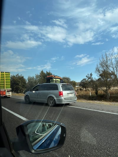 A silver minivan laden with luggage on its roof is on a sunny highway. The car is in the right lane, and its license plate is visible. A side-view mirror of the car in which the photo was taken is in the foreground. A truck with green cargo follows behind the van. The sky is blue with some clouds, and roadside vegetation lines the edge of the asphalt.