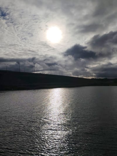 A medium-shot captures a body of water under a sky filled with mixed white and dark gray clouds, where the sun shines brightly. A bridge or overpass can be seen in the distance, supported by evenly-spaced lamp posts along its length. The lighting conditions suggest a cloudy day with the sun trying to break through. The water surface reflects the sunlight, creating a shimmering effect. The location is Нифантово, Russia.