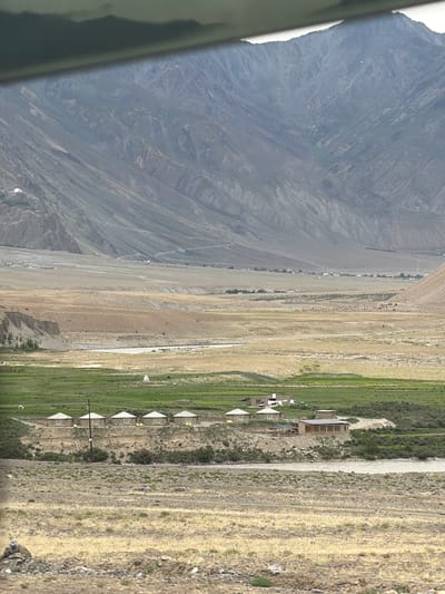 The image depicts a vast, mountainous landscape in Zangla, India. In the foreground, dry, yellowing grass covers the ground, with a rocky stream running through it. Behind the stream, a line of small, conical-roofed structures, possibly guesthouses or huts, is visible. Further back, a more substantial building with a flat roof and large windows is situated near the stream, possibly a lodge or restaurant. Green fields extend beyond these structures, with a river winding through the middle ground. The background is dominated by immense, barren, and rocky mountains, their slopes etched with the marks of time and erosion. The sky above is a muted gray, suggesting an overcast day. There are no people visible in the image.