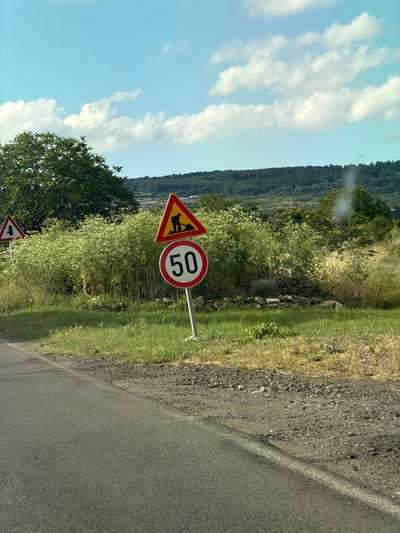 The image captures a roadside scene with two traffic signs. The prominent sign on the right is circular with a red border and white background, displaying the number "50" in black, indicating a speed limit of 50 kilometers per hour. To its left and slightly higher is a triangular warning sign with a yellow background and a black border, depicting a silhouette of a person digging. Further to the left, a smaller triangular sign with a white background and red border is partially visible. The signs are situated amidst lush green vegetation, including tall, flowering plants, and a few trees. A paved road is visible in the foreground, with a gravelly shoulder. The background features a densely wooded hill under a bright blue sky with scattered white clouds. The overall impression is of a rural or semi-rural setting, likely in or near the city of Aytos, Bulgaria, given the specific context provided. There are no people or discernible actions taking place.