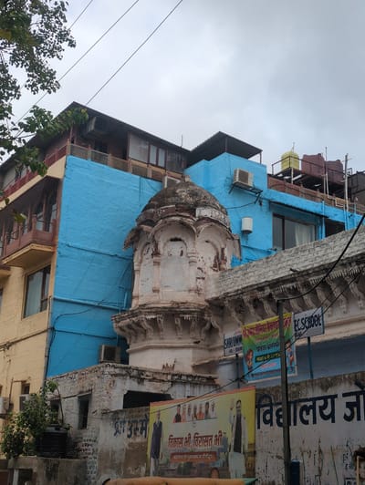 This is an exterior, eye-level, medium shot captured on an overcast day in Jodhpur, India, focusing on the exterior of buildings with a mix of architectural styles. The foreground features a textured, weathered white wall with decorative carvings along its top edge. Below this, a large banner displays images of people and text in Hindi, alongside another banner advertising "EC SCHOOL". A vertical pole partially obscures the banners and extends upwards.
Dominating the mid-ground is a light blue painted wall, appearing to be a modern addition or renovation, with an air conditioning unit mounted on it. Adjacent to this is a section of an older building painted in a pale yellow, featuring wooden balconies with intricate latticework and metal grates. A small, ancient-looking domed structure, made of weathered stone with intricate carvings, sits prominently in front of the blue wall, appearing to be part of an older, possibly religious or historical, building. Green foliage from a tree hangs from the upper left corner, with power lines crisscrossing the sky. In the background, on a rooftop, are several large water tanks, one yellow and two dark reddish-brown, suggesting the location is likely in a densely populated urban area where water storage is common. The overall impression is of a neighborhood where different eras of architecture coexist.