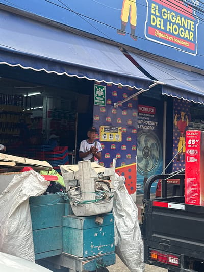A man stands with his hands clasped in front of a storefront in Perímetro Urbano Barranquilla, Colombia. The storefront features a blue awning and a large blue sign with yellow lettering that reads "EL GIGANTE DEL HOGAR" and "Gigante en economía". Next to the man is a large fan advertisement. In the foreground, various items are piled up, including sacks, blue crates, and cardboard boxes. A black pickup truck is parked to the right of the scene. The overall environment appears to be a commercial area.