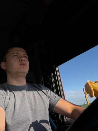 A man is driving a vehicle. He is wearing a gray t-shirt. He is looking up and to the right. A peeled banana is visible in the passenger seat. The sky is blue and there is a green landscape in the background. The location is the city of Borskoe, Russia.