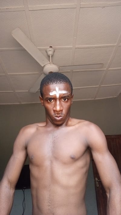 A shirtless, dark-skinned man faces the camera with a serious expression. A white cross is painted on his forehead. He has short, dark hair. Above him is a white ceiling with a white ceiling fan. A television and a door are partially visible in the background. The image is well-lit, revealing the man's physique and facial features clearly.