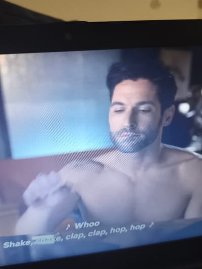 The image is a photograph of a digital screen displaying a video frame. The screen's black bezel is visible at the top and bottom.
Within the video frame, a male individual is prominently featured, positioned centrally. He is Caucasian, appears to be in his mid-30s to early 40s, with short dark hair and a neatly trimmed beard. He is shirtless, revealing a muscular upper torso and shoulders. His head is tilted slightly down, and his gaze is directed downwards and to the left. His right arm is raised and bent, with a light-colored, amorphous object near his right shoulder/bicep area, possibly a prop or part of an action.
The background behind the male is out of focus. To his left, a light blue glow emanates, and a reddish-brown, blurred object, potentially furniture, is partially visible. The right side of the background is darker and indistinct.
Overlaying the bottom of the screen content is white text with musical notation: "♫ Whoo Shake, shake, clap, clap, hop, hop ♫". The word "Shake" is partially obscured.
The location depicted within the screen's content is an indoor set. There are no identifiable features within this image to determine a specific real-world location, including any location within the city of Panicheri, Bulgaria. The image portrays fictional media content, not a live event in a specified geographic location.