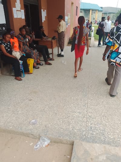 The scene depicts an outdoor area adjacent to a reddish-brown building in Rumuekini, Nigeria, likely an administrative or academic facility, with a multi-story building visible in the background.
In the foreground, a concrete and tiled walkway shows scattered clear plastic refuse. To the left, five individuals are seated on a low bench or ledge against the building wall. From left to right: a female in a black logo-patterned top and black pants holding a white bundle; a female in an orange top with sunglasses pushed onto her head; a female in a colorful patterned dress; a male in a black shirt actively using a mobile phone; and another person in black, partially obscured. A yellow plastic bag rests on the ground near the seated group.
Further right, a person in a light brown uniform, dark pants, black cap, and black shoes with prominent white socks stands behind a wooden table positioned against the building wall. White papers are taped to the wall directly behind this area.
In the mid-ground, a female with long, braided reddish-brown hair, wearing a red skirt and a colorful patterned sleeveless top, carrying a black bag, walks away from the viewer. To her right, a male in a colorful geometric patterned shirt and light brown pants is also walking away. Several other individuals are visible in the distant background, indicating a populated area. The sky is overcast.