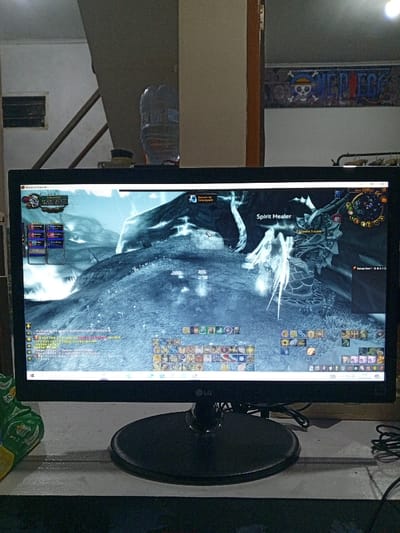 The image displays an indoor scene, specifically a computer workstation located in Al Haouafate, Morocco. A black LG computer monitor, resting on a light-colored desk, dominates the foreground. The monitor is actively displaying the video game "World of Warcraft," featuring a snowy, mountainous virtual environment. The game interface shows a "Spirit Healer" character and a "Return to Graveyard" message, indicating the player's in-game character is currently incapacitated. Spell icons, a mini-map, and chat logs are visible elements of the user interface. To the left of the monitor, a partially visible green and yellow snack bag rests on the desk. To the right, black cables are coiled. In the background, behind the monitor, a multi-level interior is visible. On the left, a section of a staircase with light-colored walls and wooden steps is present, along with a rectangular vent opening and a clear plastic water bottle. On the right, a white wall features a "One Piece" anime banner displaying a skull and crossbones with the words "ONE PIECE" and "MEP FGC". A light source is partially visible at the top right of the background.