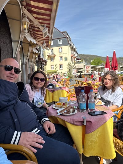 The image depicts three individuals seated at a table in an outdoor café in Cochem, Germany. The setting is a tourist area, evident from the architecture and the presence of awnings. The table is covered with a yellow and pink tablecloth, with water bottles, menus, a smartphone, and plates of food (cake, pastries) placed on it. The first person, likely a male, has a bald head and wears sunglasses and a navy jacket with white stripes on the sleeves. The second person, a female, wears sunglasses, a white t-shirt, and a light pink jacket. The third person, up front, has curly hair and a gray shirt. They appear to be relaxing and possibly enjoying a meal. The background reveals a street scene with other people and buildings typical of Cochem's architecture.
