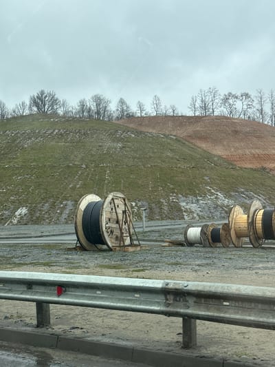 The image shows a roadside scene in what appears to be a rural area. The sky is overcast. Along the side of the road, to the right of the frame, there's a metal guardrail. Next to the guardrail are large wooden cable spools containing various types of cables or wires. The cables are in various colors. In the background, there is a grassy slope leading up to a barren, reddish-brown hill. Leafless trees are visible on the hillside. The ground appears to be wet and muddy. There are no people visible in the image. The location is specified as Grebeni, Russia.