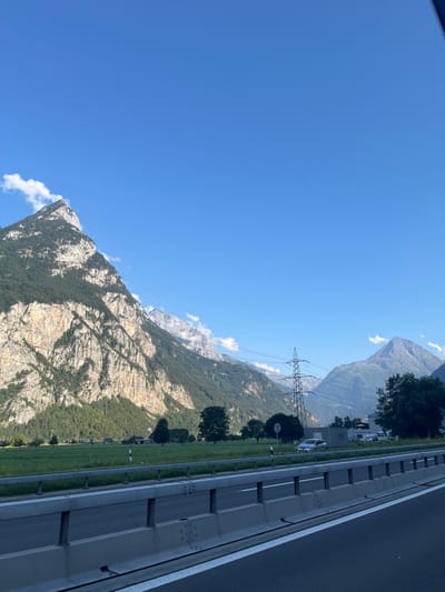 The image presents a daytime outdoor scene captured from the perspective of a vehicle on a multi-lane highway, located on the A2 highway near Schattdorf, Canton Uri, Switzerland. In the foreground, a concrete highway barrier with a metal guardrail extends from left to right, above which asphalt roadway lanes are visible. Beyond the barrier, a flat, expansive green grassy field occupies the left midground, dotted with scattered deciduous trees. A light-colored sedan is parked on the grassy shoulder alongside the highway, positioned behind the guardrail. A prominent, tall, grey metal lattice electricity pylon stands in the central-right midground, supporting overhead power lines that stretch horizontally across the frame. Further to the right, an area featuring more trees and indistinct buildings is present. The background is dominated by large, steep mountains under a clear, bright blue sky with minimal cloud cover. The most prominent mountain, on the left, features a distinctive light-colored, sunlit rock face with dense green forestation covering its upper and lower slopes, and a small white cloud is visible near its peak. Other mountains recede into the distance on the right, some partially obscured by atmospheric haze. No people, specific actions, or interactions are discernible within the scene.