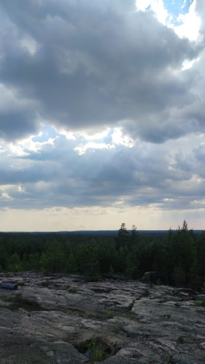 The image depicts a vast, rocky expanse in the foreground, leading to a dense, dark green forest that stretches across the horizon. Above the forest, a hazy, pale sky is visible, partially obscured by dramatic, thick clouds. A faint plume of smoke is seen rising from the distant treeline on the left side of the frame. The overall impression is a natural, rugged landscape under an overcast sky. This scene is located in Pitkyaranta, Russia, a region known for its forests and rocky terrain.