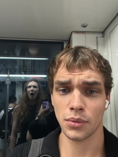 A young man takes a selfie on a subway. He is in the foreground, with his face taking up most of the frame. He has brown hair, blue eyes, and is wearing a black jacket and white earbuds. Behind him, a young woman is visible in the reflection of the subway window. She has long brown hair and is looking at the camera with her mouth open in surprise. There are other passengers in the background, blurred and indistinct. The subway car appears to be modern and clean. The overall impression is a candid shot capturing a moment of surprise on a commute.