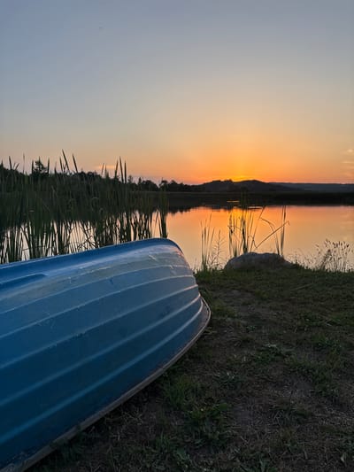 An upside-down blue rowboat rests on a grassy bank beside a calm body of water. Tall reeds line the water's edge, reflecting the warm hues of a sunset. In the distance, rolling hills are silhouetted against the orange and yellow sky. The scene appears to be a natural setting, possibly a lake or pond, within or near Skalsko, Bulgaria. There are no people visible in the image.
