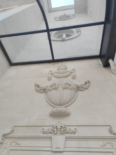 The image depicts a section of an ornate, light-colored building facade under a modern glass and metal roof structure. The facade features classical-style decorative elements, including a large, sculpted vase motif with floral details and scrolls, positioned above a decorative frieze or panel with additional scrollwork and a centered shield-like element. Above the vase, there appears to be another decorative element with a garland or swag. The glass roof structure consists of multiple panes divided by a black metal frame. Visible through the glass roof are higher portions of the building, including round windows with white frames. The location is identified as George, South Africa, although specific locational details within the city are not discernible from the image. No people or specific actions are observable.