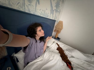 A young woman with dark, curly hair is sitting on a bed in a bedroom. She is wearing a light purple t-shirt and looking upwards with her left hand extended. The bed is covered in white linens. To the right of the woman, a small, reddish-brown dog toy is lying on the bed. In the background, a dark blue headboard is visible, and the wall behind it is decorated with wallpaper featuring a blue and white toile pattern with giraffes and foliage. A bedside table with a woven lamp is situated next to the headboard. The image does not contain any identifiable landmarks or features that would place it in the city of Banya, Bulgaria.