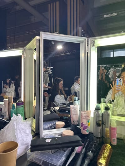 The image depicts an indoor scene within a preparation or dressing area, likely located in Issy-les-Moulineaux, France. The foreground presents a cluttered vanity surface, viewed from above, covered with numerous beauty and hair products. Visible items include multiple bottles of hairspray and styling creams (brands like Kérasoin "Easy Curls" and "Silky"), various hairbrushes (round and flat), a hair dryer, a hair straightener, a black brush roll, several small cosmetic containers, a crumpled white paper, and a brown paper cup.

Central to the image is a three-panel vanity mirror unit with a silver frame, equipped with integrated fluorescent lights on its side panels. The reflections within these mirrors reveal several individuals and the surrounding environment.

In the central mirror, a female subject with dark hair, wearing a black and white garment, is seen having her hair styled by an individual whose hands, holding a heated hair tool, are visible. The subject is looking down at a smartphone. To her left, reflected in the left mirror panel, another female subject with dark hair is visible, wearing a white robe and also engaged with a smartphone. To the right of the central subject, a partially visible individual in a white garment is present.

The rightmost mirror reflects the person taking the photograph; only their upper face, hands, and a smartphone with a colorful, possibly iridescent, case are visible. This individual is wearing a white robe.

The background, visible both directly and in reflection, reveals a dark, expansive interior space, indicative of a studio or event hall. Overhead, industrial elements such as exposed ducts and lighting fixtures are apparent. Vertical striped pillars (black and light brown) stand in the background. Clothing racks holding various garments, including what appear to be bras and other apparel, are visible, suggesting a wardrobe area adjacent to the preparation space. Bright, square light sources are also reflected in the distance.