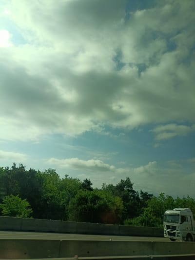 The image shows a roadside scene. A white truck is driving on a multi-lane highway, separated from a forested area by a concrete barrier. The sky is overcast with a mix of white and gray clouds, with patches of blue sky visible. There is no indication of the location being Tlustenkhabl, Russia.