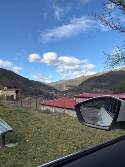 The image presents a panoramic view of a mountainous valley landscape in the city of Tompsan, Bulgaria, observed from the interior of a vehicle. The immediate foreground, at the bottom right, reveals the dark, textured interior door panel of a car and a silver side-view mirror. The mirror's reflection shows the side of the vehicle, a section of paved ground, and a building with a red tile roof. To the lower left, a segment of a weathered wooden picnic bench, painted blue with visible paint loss, rests on a sparse green and brown grassy area.

In the midground, a rudimentary fence composed of metal posts and wires traverses the scene. To the left of this fence stands a derelict, multi-level concrete building exhibiting structural damage, exposed rebar, and missing roof sections. A railway line or narrow road curves through the valley, bordered by utility poles or communication lines. Several buildings with prominent red tile roofs are scattered across the valley floor and lower hillsides, indicating a residential settlement.

The background features a series of undulating mountains and hills, predominantly covered with a mix of evergreen and deciduous trees, many of which are bare or show initial signs of budding. Patches of snow are discernible on the higher elevations. The sky is clear blue with scattered white cumulus clouds. Bare tree branches extend into the upper right quadrant of the image. No individuals or active interactions are visible.