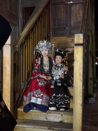 Two individuals are positioned on wooden steps within what appears to be an enclosed, traditional structure. An adult female, adorned in elaborate traditional attire including a large silver headdress, heavy necklaces, and a red patterned robe, is lying on the steps next to a child dressed in a black costume with silver embellishments and a distinctive tall, curved hat. The structure features aged wooden planks and beams, with dark wooden panels on the left. The setting suggests a cultural performance or a staged event taking place in a historic area or building. The individuals are lying, not standing or sitting, on the steps. There is no definitive information in the image to confirm its location in Zhuoshui, China, but the traditional clothing suggests a region with distinct cultural customs.